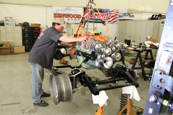 man lowering a v8 engine onto a vintage hot rod vehicle chassis