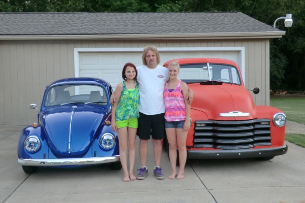 a man stands with his daughters near a vintage chevy 3100 truck and vw beetle bug