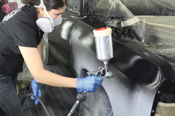 woman spraying black paint onto an old truck fender in a spray booth