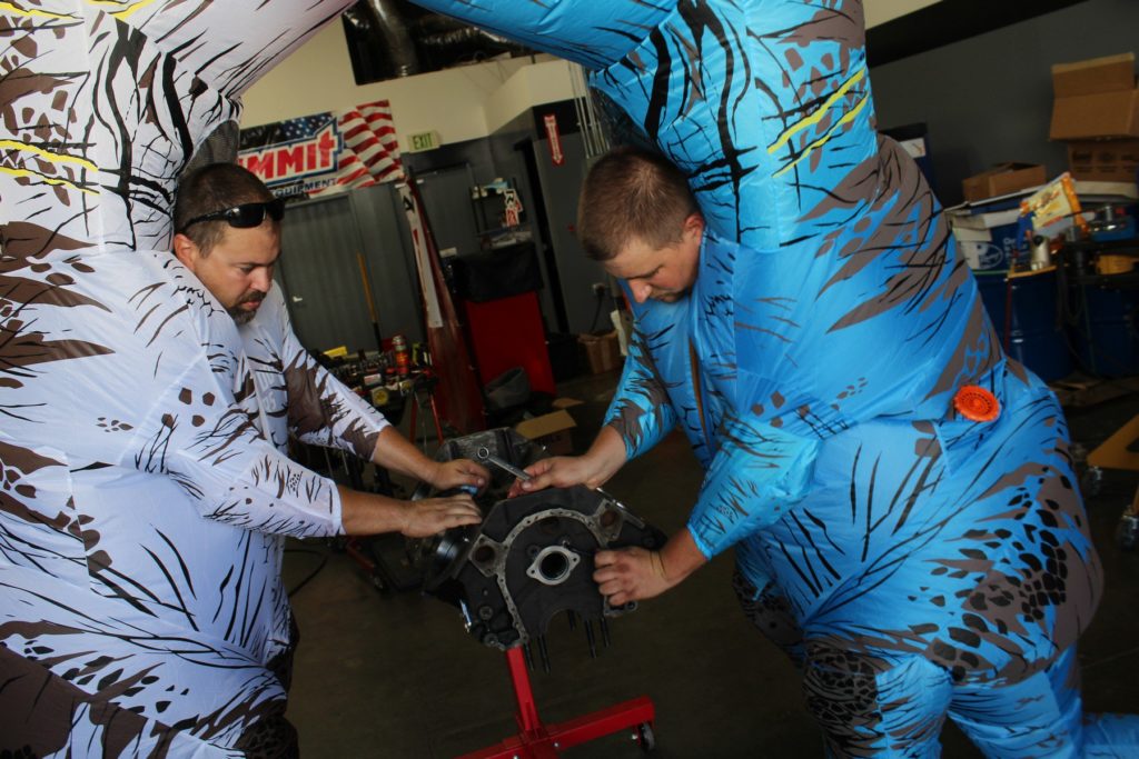 men in dinosaur costumes disassemble an engine block