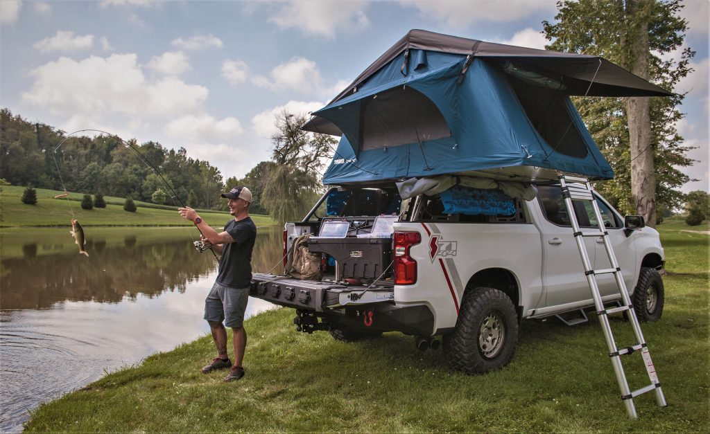 a guy fishing next to an off road overlanding modified chevy Silverado truck