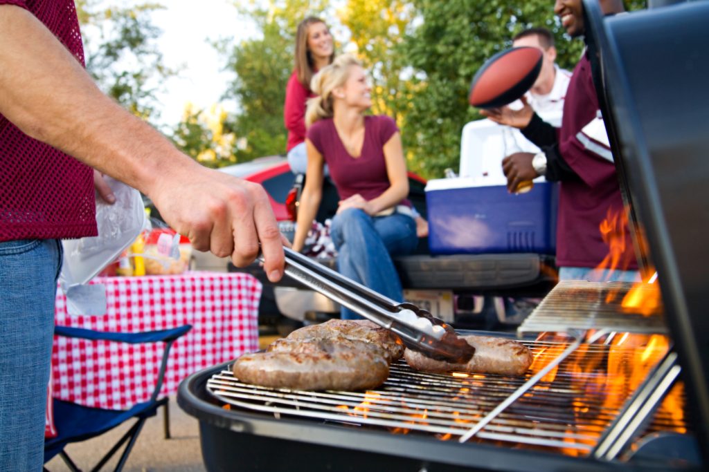 group of people enjoyin a tailgate party with grill and burgers in foreground