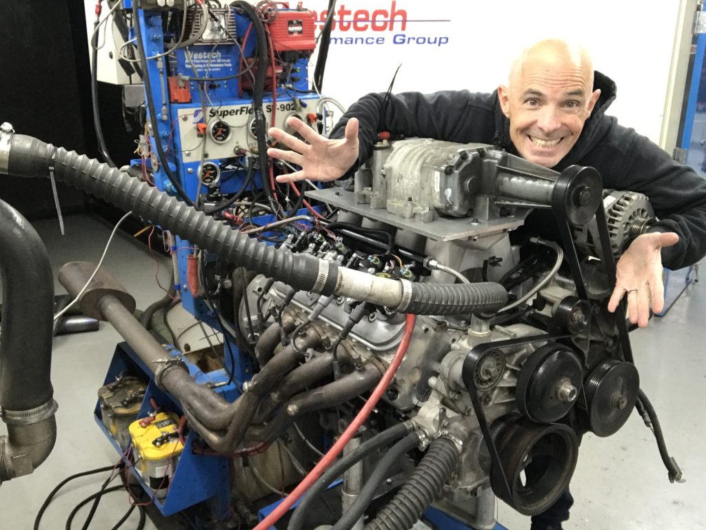 man posing next to a supercharged engine on a dyno